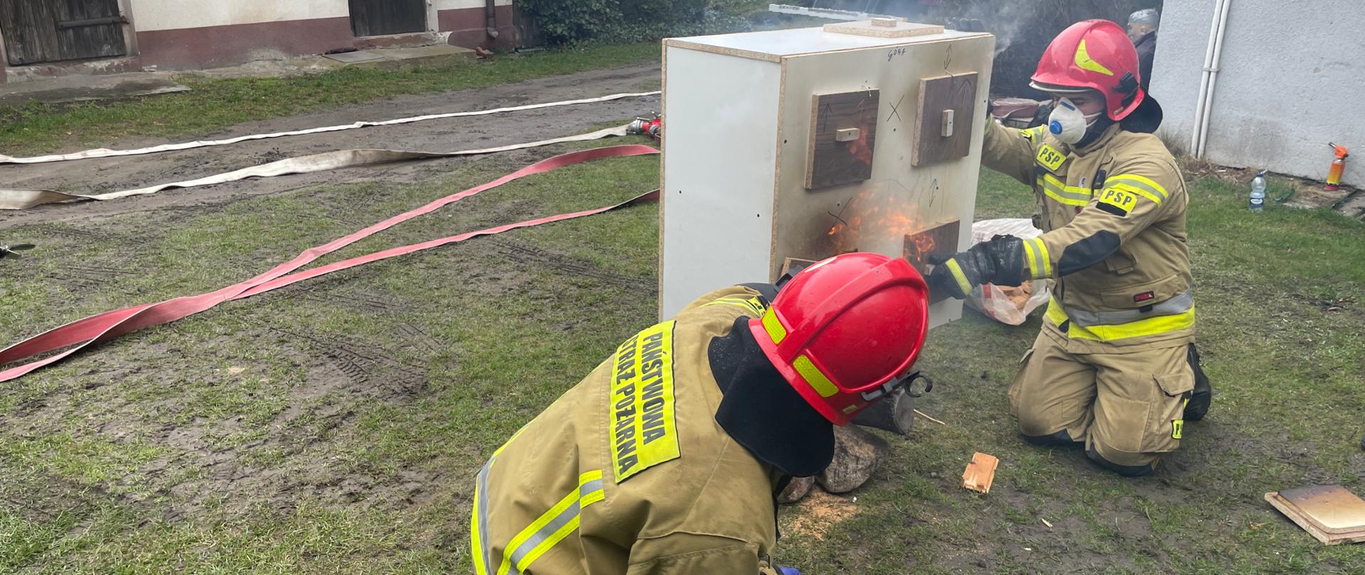 dwóch strażaków klęczy i prezentuje na podstawie modelu domu pożar, wo oddali budynki i samochód strażacki 
