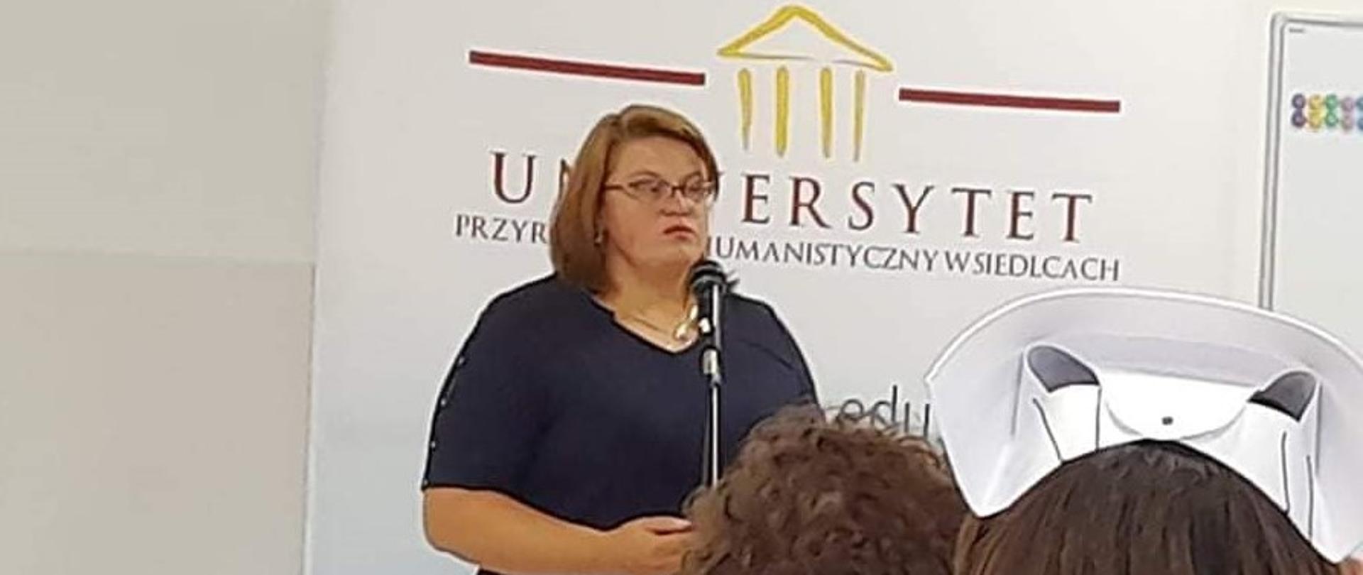 Na zdjęciu znajduje się kobieta przemawiająca do mikrofonu