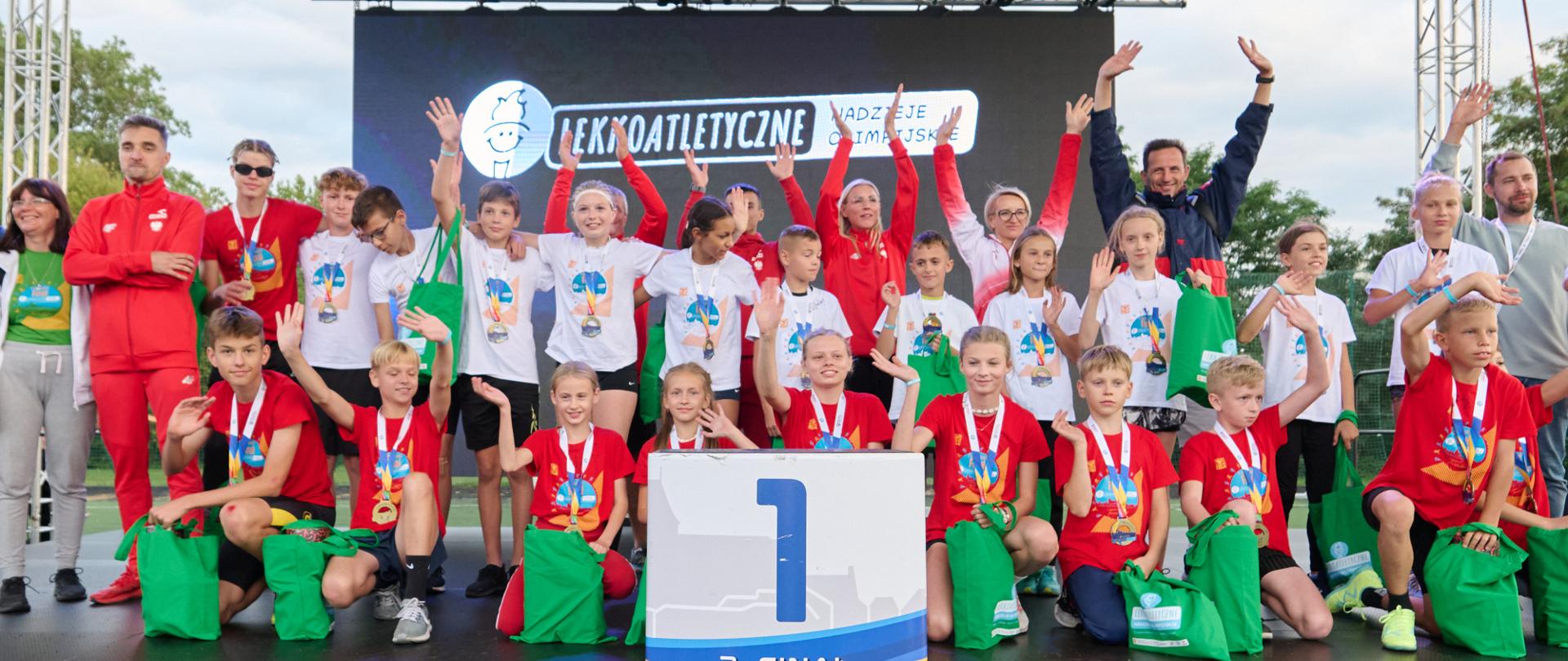 Pod daszkiem stoi i siedzi duża grupa młodzieży w białych i czerwonych koszulkach, machają rękami, przy nich leżą zielone worki, przed nimi rozpostarty plakat z wielką jedynką.