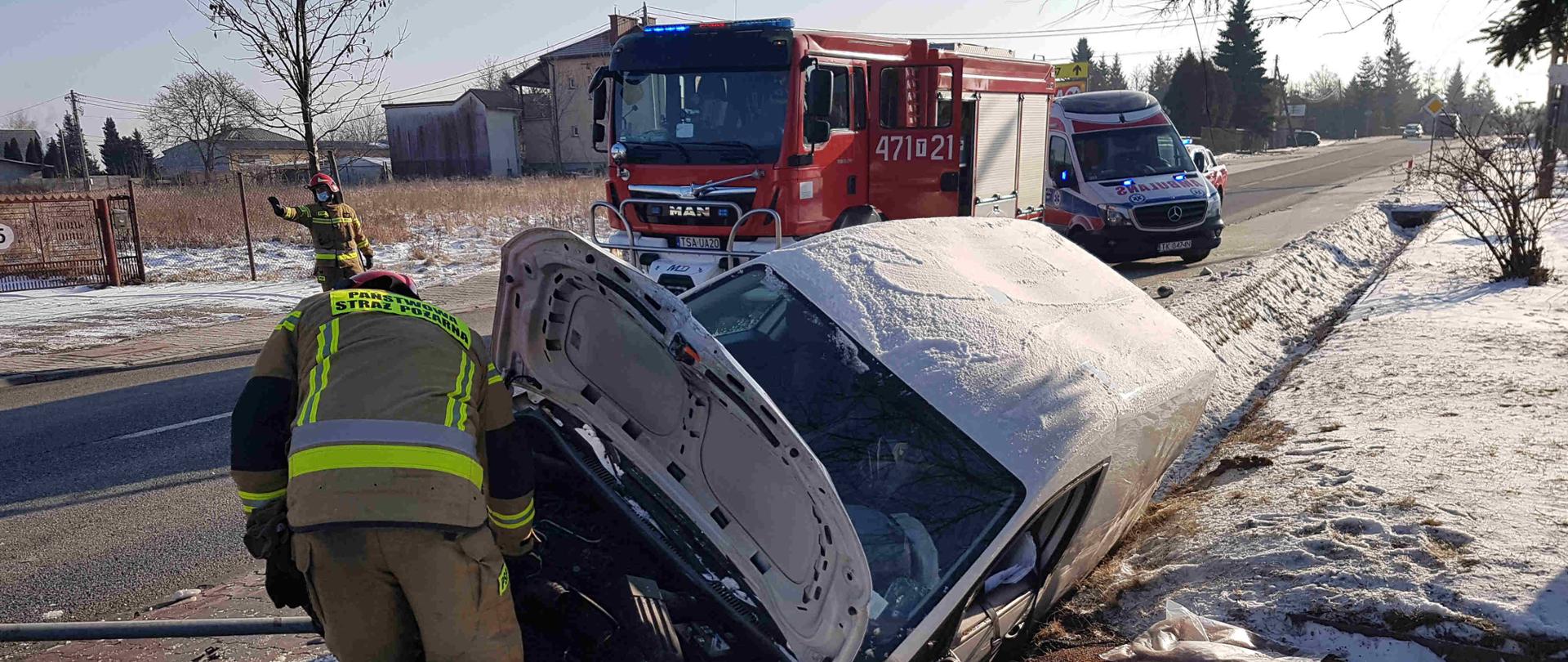Zdjęcie zrobione w dzień na drodze. Na zdjęciu widać rozbity samochód znajdujący się w rowie i strażaków oraz samochód strażacki, karetkę pogotowia.