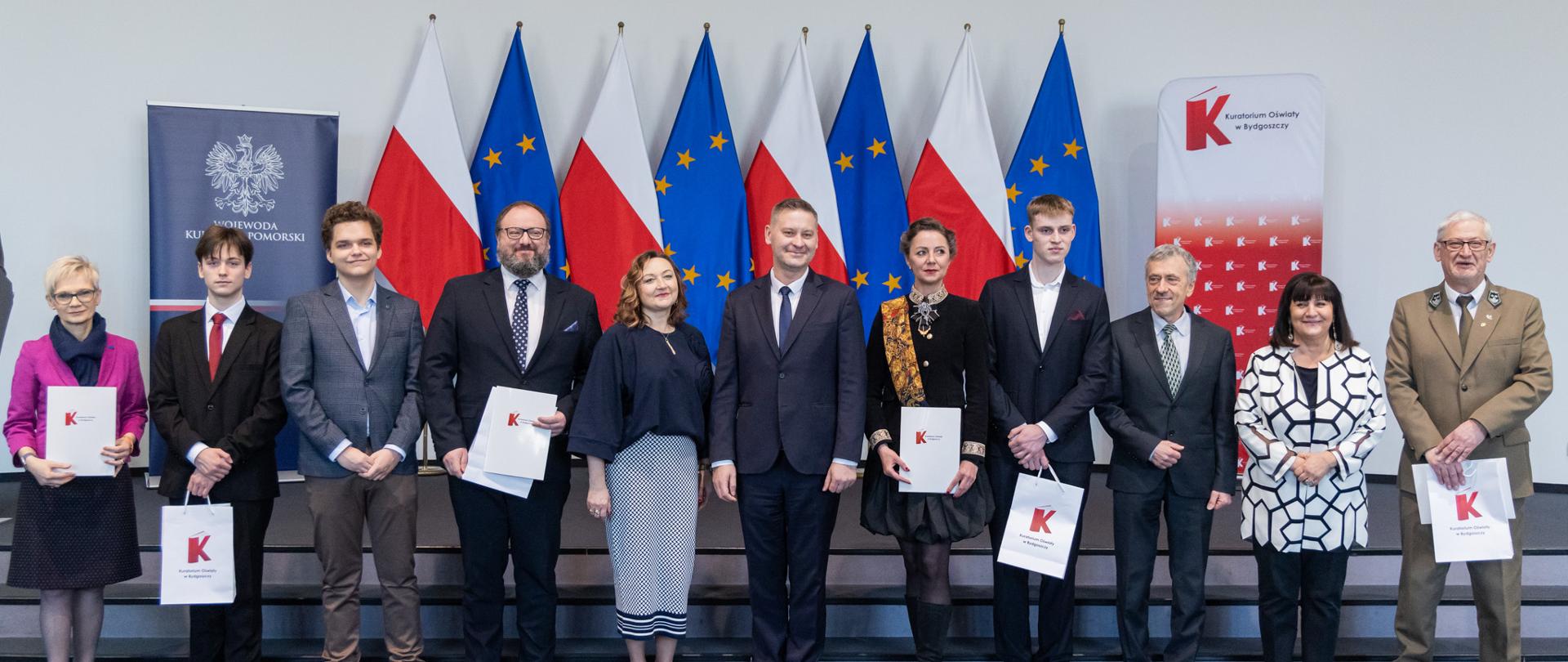 Grupowe zdjęcie pamiątkowe na scenie podczas Gali Rankingu Perspektywy 2026 „Najlepsze szkoły regionu”. W centrum grupy stoi wicewojewoda Michał Koniuch w ciemnym garniturze oraz Kujawsko-Pomorski Kurator Oświaty Grażyna Dziedzic, otoczeni są przez nagrodzonych przedstawicieli szkół trzymających nagrody. W tle, za uczestnikami, widoczne są oficjalne flagi (polski oraz unijna). Wszyscy są uśmiechnięci. 