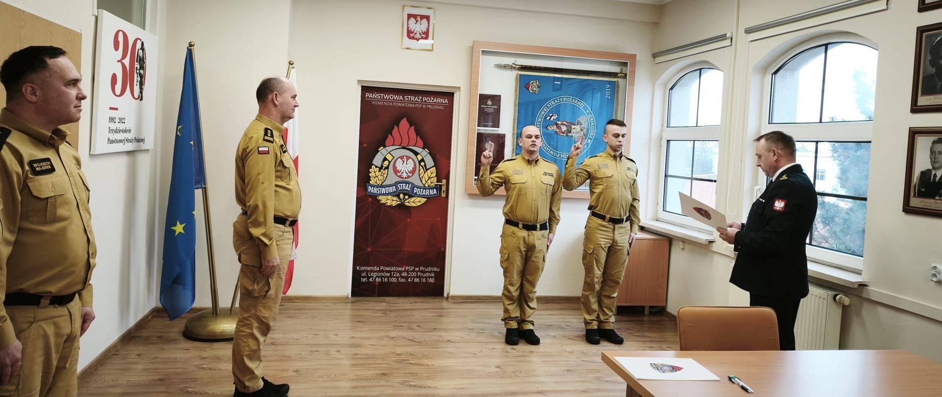 czterech strażaków Państwowej Straży Pożarnej stoi w sali komendy PSP w Prudniku podczas uroczystości służbowej; dwóch w jasnych mundurach trzyma dokumenty, obok nich oficer w galowym mundurze, w tle flagi, godło Polski i sztandar