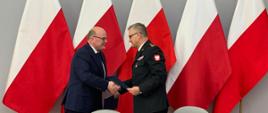 Za stołem prezydialnym stoją komendant wojewódzki i prezydent miasta Grudziądza. Ściskają sobie dłonie i wymieniają podpisane dokumenty. W tle flagi narodowe w szeregu.