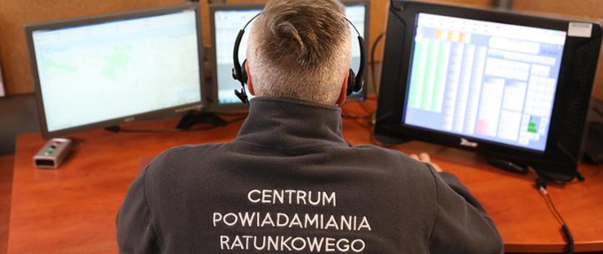 Zdjęcie przedstawia operatora numeru alarmowego 112. Widok na plecy i monitory