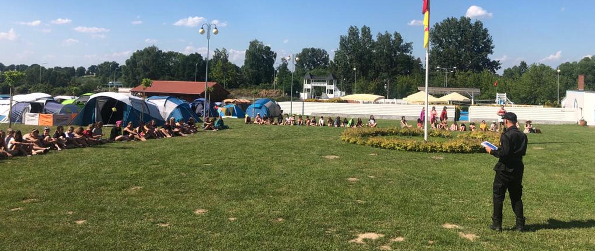 Na zdjęciu funkcjonariusz podczas pogadanki z młodzieżą nt. bezpieczeństwa na środku masz z flagą obozu oraz namioty i domki miejsca wypoczynku