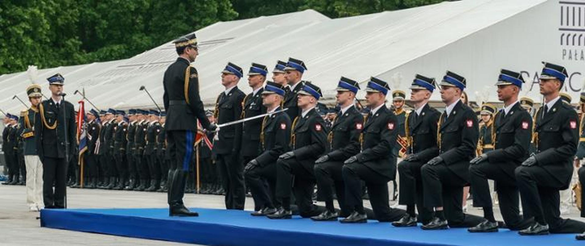Szereg strażaków w mundurze galowym, część z nich klęczy na jednym kolanie, strażak stojący dotyka ramienia strażaka klęczącego szablą. W tle pododdział strażaków, namioty. 
