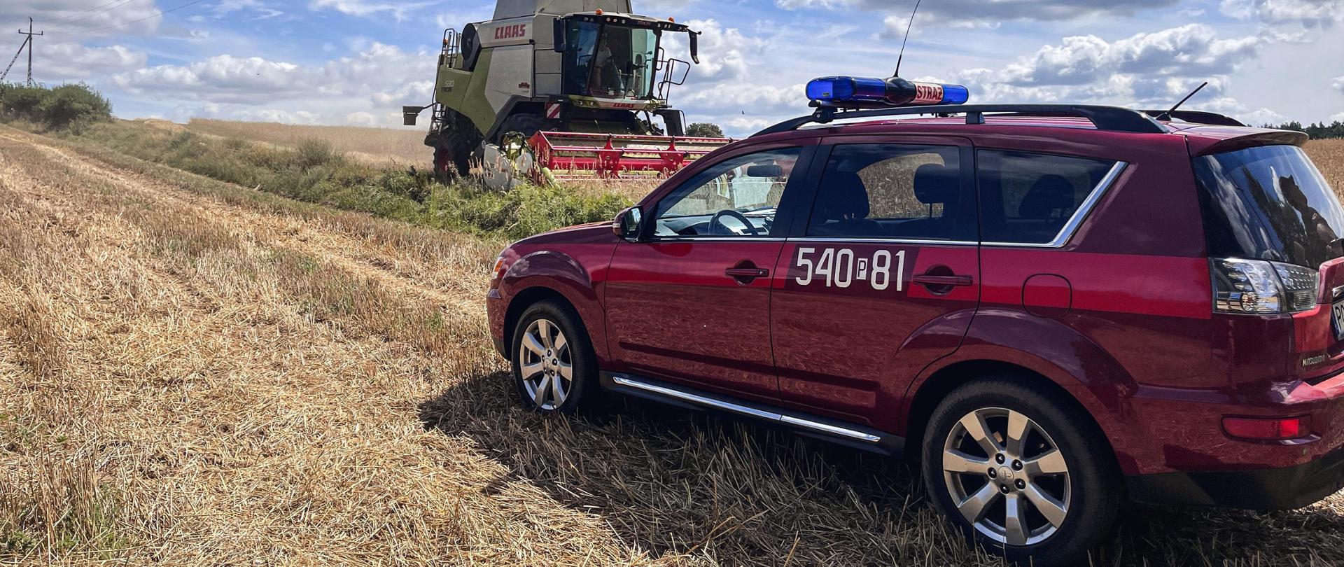 Samochód operacyjny straży pożarnej stoi na polu. Na drugim planie trwa młócenie zboża.