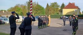 Uroczystość z okazji Dnia Flagi Rzeczypospolitej Polskiej. Strażacy ze zmian służbowych stoją naprzeciwko siebie. Poczet flagowy wciąga flagę państwową na maszt. Komendant, Zastępca Komendanta, poczet flagowy i dowódcy zmian salutują.