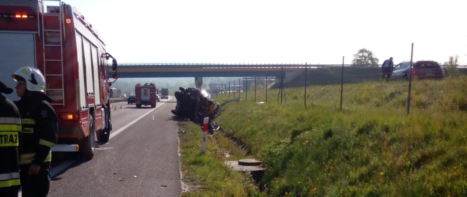 Zdjęcie przedstawia drogę S7. Po prawej stronie drogi, w przydrożnym rowie, na boku leży samochód dostawczy, który uległ wypadkowi. Po lewej, na pasie jezdni stoją dwa wozy bojowe. Na pierwszym planie widać dwóch ratowników.