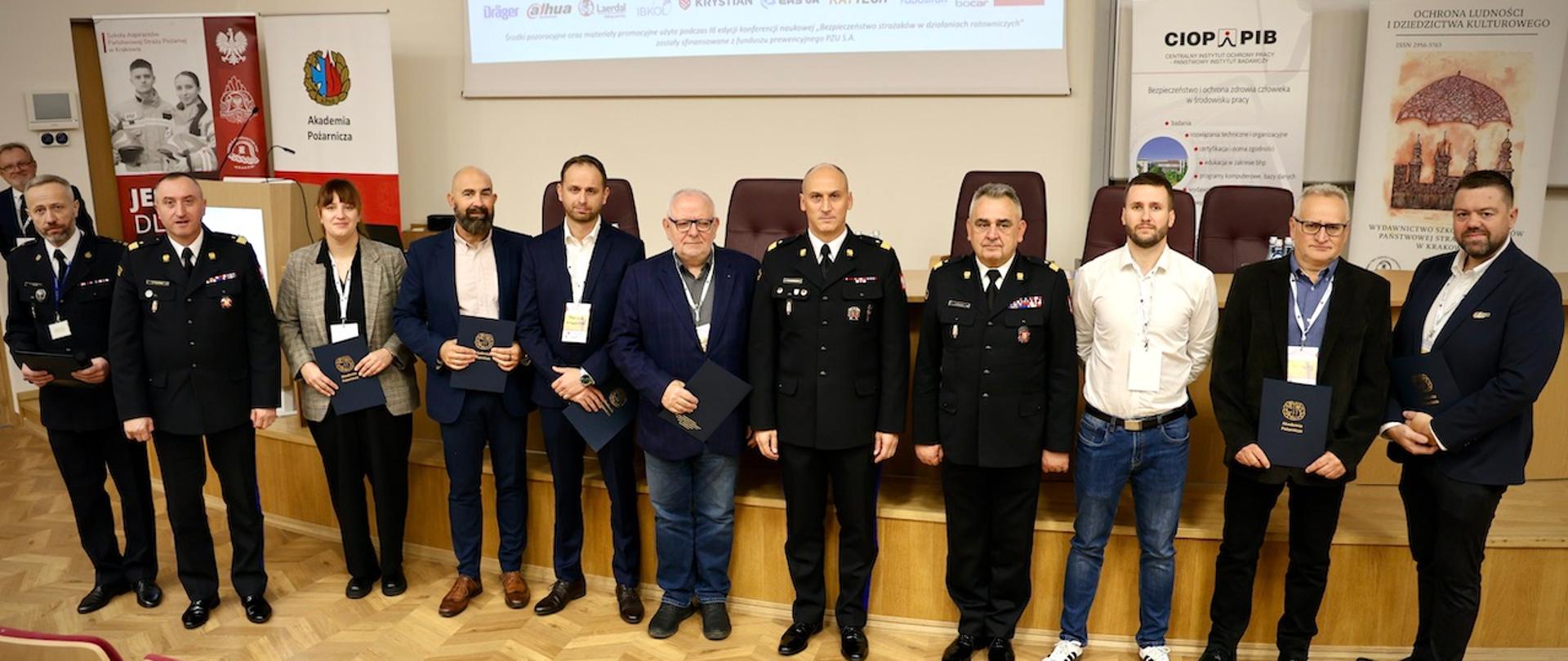 Grupa uczestników konferencji, w tym funkcjonariusze PSP w mundurach wyjściowych i przedstawiciele instytucji partnerskich, stoi na scenie w sali wykładowej. W tle banery Akademii Pożarniczej i partnerów wydarzenia.