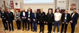 Grupa uczestników konferencji, w tym funkcjonariusze PSP w mundurach wyjściowych i przedstawiciele instytucji partnerskich, stoi na scenie w sali wykładowej. W tle banery Akademii Pożarniczej i partnerów wydarzenia.