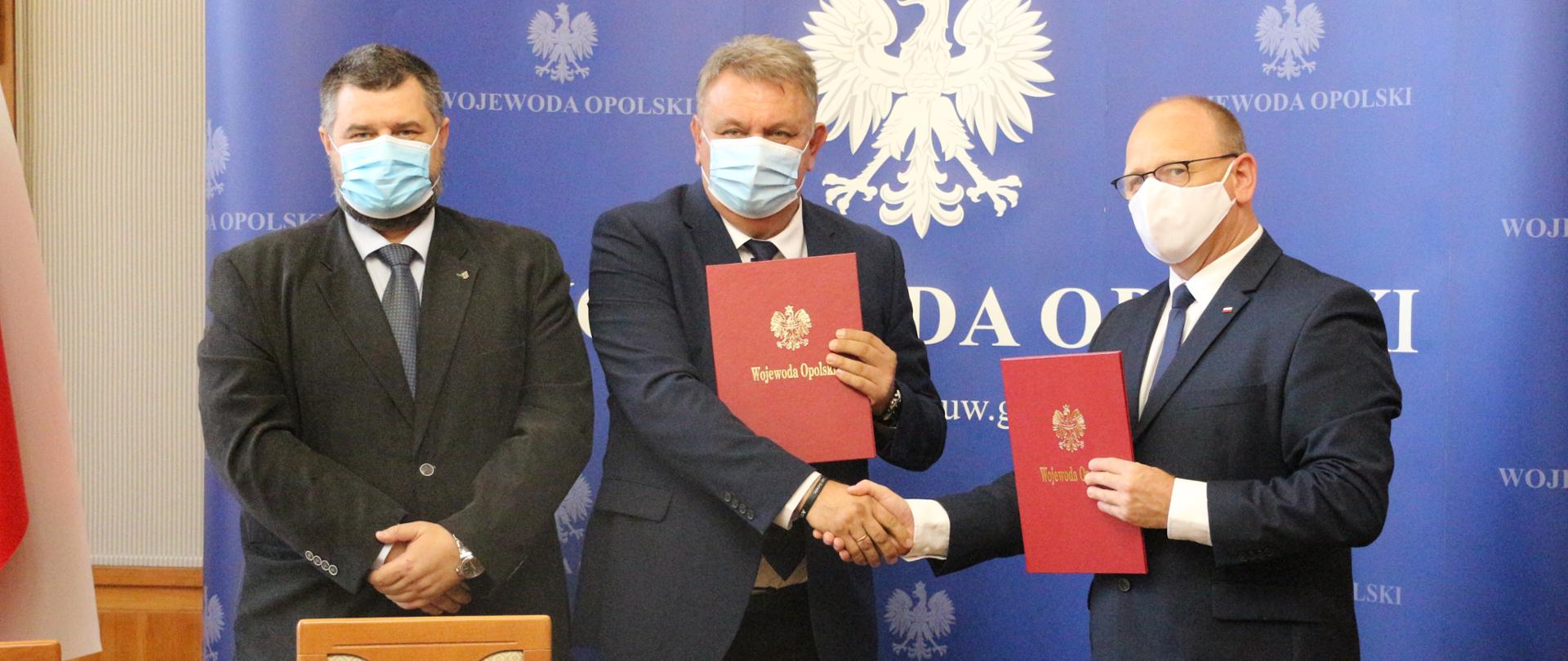Od lewej strony dr Bartosz Kuświk, dyrektor Instytutu Śląskiego, Dariusz Byczkowski, dyrektor Muzeum Piastów Śląskich w Brzegu oraz Mariusz Nieckarz, dyrektor Biura Wojewody opolskiego i pełnomocnik Wojewody Opolskiego ds. Programu "Niepodległa".. Mężczyźni stoją na tle granatowej ścianki z napisem Wojewoda Opolski. Dariusz Byczkowski pozostaje w uścisku dłoni z Mariuszem Nieckarzem. W swoich dłoniach trzymają także teczki z napisem Wojewoda Opolski. Przed nimi znajduje się blat drewnianego, okrągłego stołu. Na nim dwie pieczątki i długopis. Po lewej stronie kadru widać fragment biało-czerwonej flagi. Mężczyźni zasłaniają usta i nosa za pomocą maseczek ochronnych. 