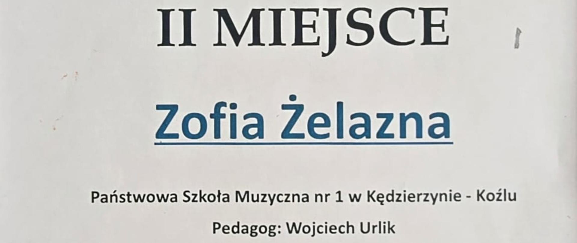 Na plakacie w jasnej, białej kolorystyce widoczny jest dyplom z niebieskimi i czarnymi elementami graficznymi, w tym logo z kluczem wiolinowym i postacią dziecka oraz ozdobnym motywem fal na dole.
Centralnie dużą czcionką zapisano „II MIEJSCE”, poniżej imię i nazwisko „Zofia Żelazna”, a wyżej informację o VI Ogólnopolskim Konkursie „Co Młodym w Duszy Gra?” wraz z datą 23–25 kwietnia 2026 roku i miejscem wydarzenia.
W dolnej części znajdują się nazwa szkoły, nazwisko pedagoga, podpisy jury i dyrektora oraz rząd logotypów patronów i sponsorów.