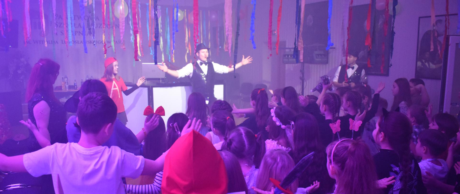 na zdjęciu, w przystrojonej karnawałowo auli PSM widnieją uczniowie, którzy powtarzają za wodzirejem ruchy taneczne. Kolorystyka zdjęcia jest biało-żółto- czerwono-niebiesko-czarna. 
