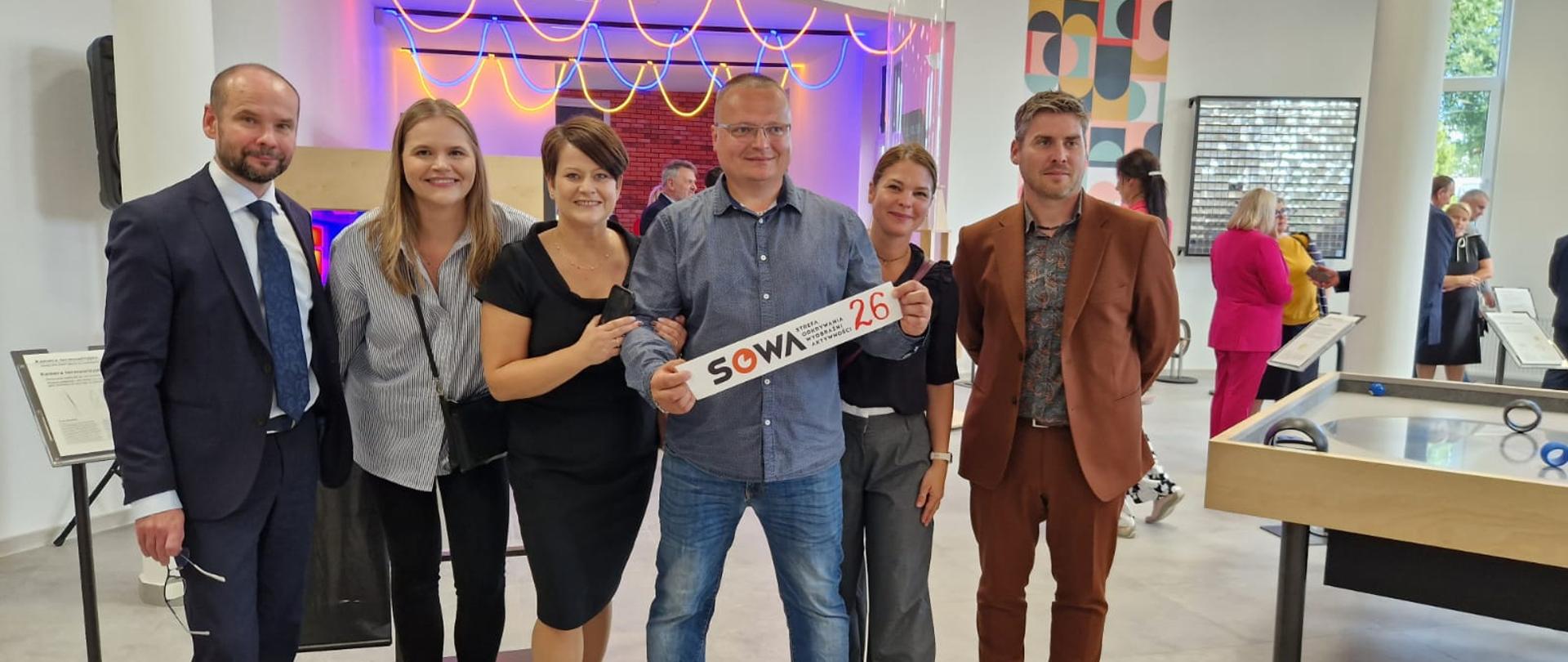W sali na tle oświetlonej na kolorowo ściany stoi grupa kilku osób, mężczyzna w szarej koszuli z przodu trzyma małą tabliczkę z napisem SOWA.