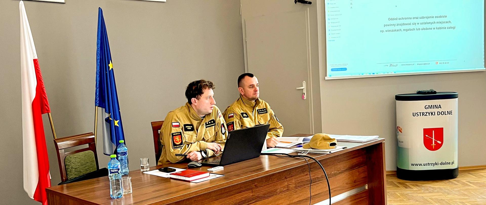 Zdjęcie przedstawia prowadzących szkolenie oficerów PSP siedzących przy biurku. W tle włączona prezentacja. 