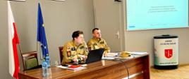 Zdjęcie przedstawia prowadzących szkolenie oficerów PSP siedzących przy biurku. W tle włączona prezentacja. 