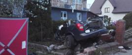 Całkowicie zniszczony samochód osobowy stojący na posesji przy drodze. W aucie obcięto dach i drzwi od strony kierowcy. Widoczne zniszczone ogrodzenie. Obok rozstawiony strażacki parawan.