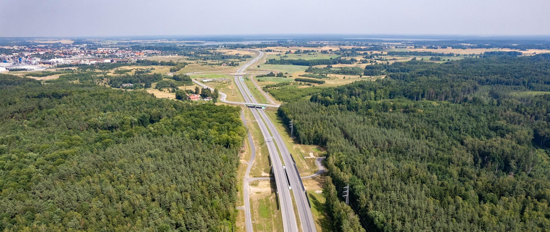 Zdjęcie lotnicze przedstawiające dwujezdniową drogę ekspresową biegnącą przez gęsty kompleks leśny. Po obu stronach trasy rozciągają się duże połacie lasów, a w oddali widać pola, zabudowania oraz skrzyżowanie z wiaduktem.