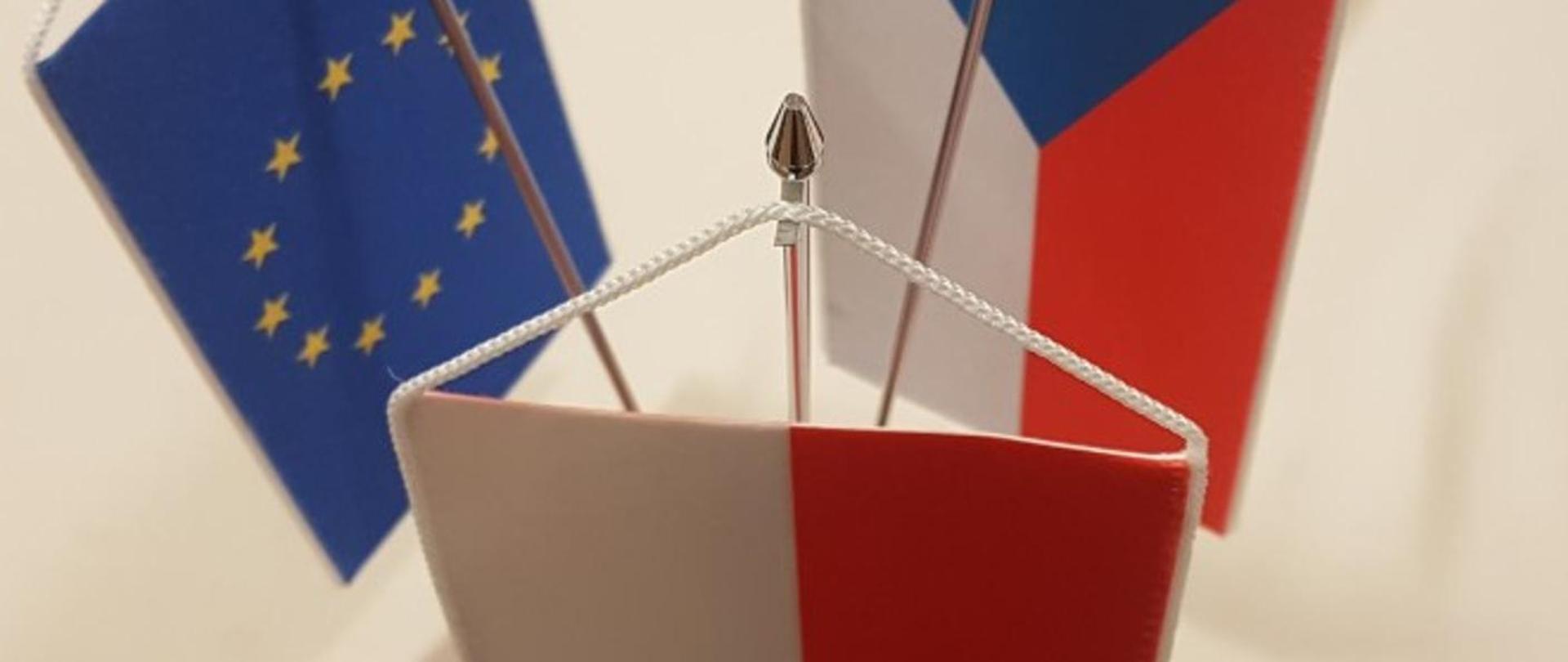 Trzy małe stojące flagi: z przodu flaga Polski, z lewej flaga Unii Europejskiej, a z prawej flaga Czech, ustawione blisko siebie na wspólnym stojaku.
