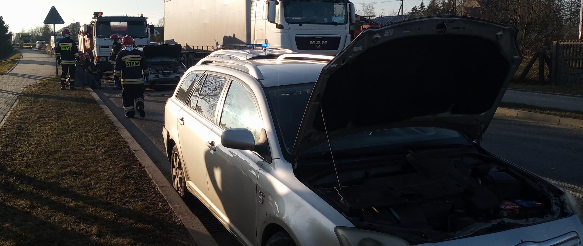 Na zdjęciu widoczny uszkodzony pojazd marki Toyota, w tle widoczni strażacy udzielający kwalifikowanej pierwszej pomocy poszkodowanej w wypadku kobiecie.