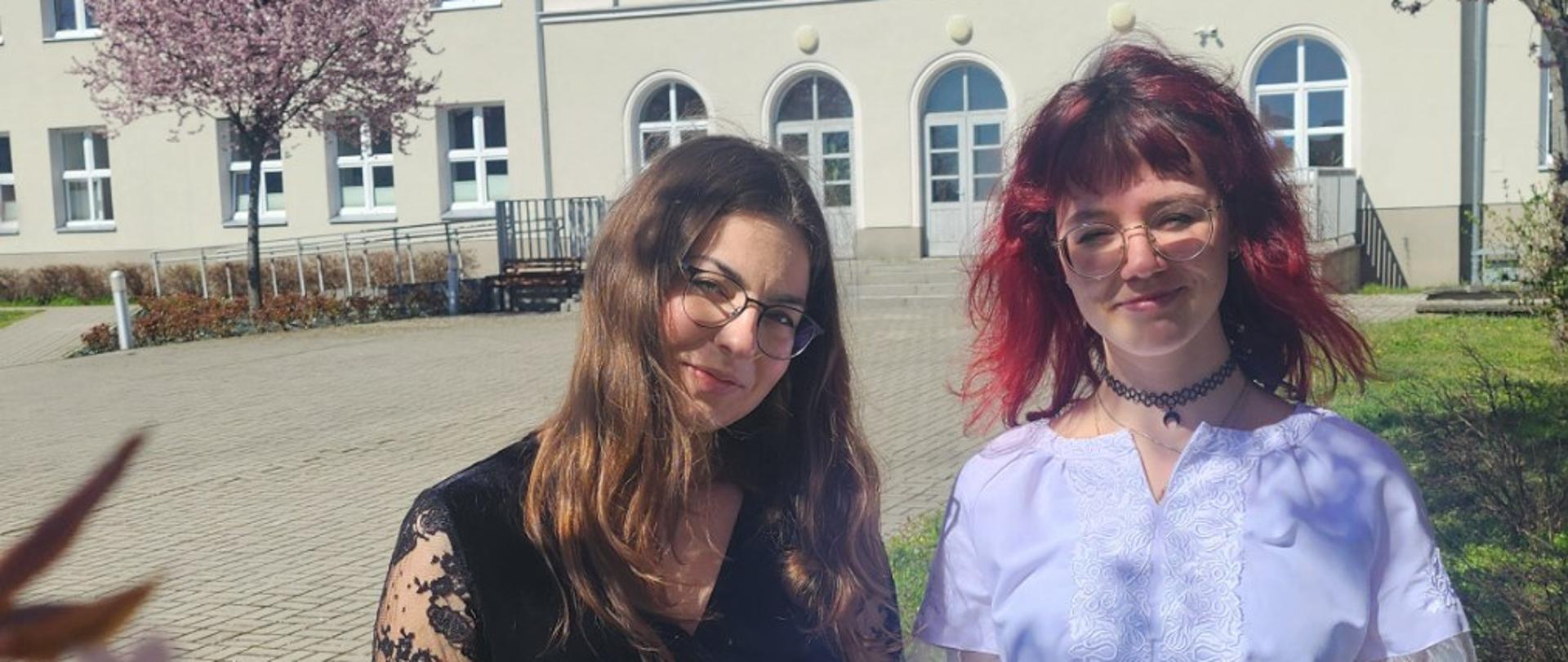 Paulina Zubel i Olga Drozdovska z dyplomami z Festiwalu Pianistycznego im. Lidii Grychtółówny. W tle budynek szkoły i kwitnące drzewa