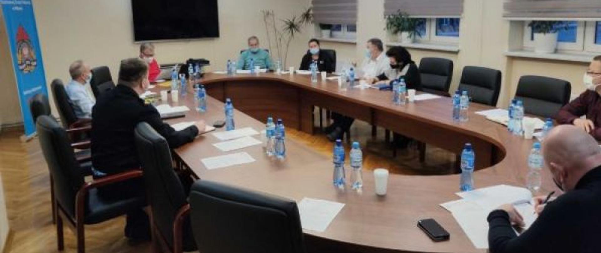 Na zdjęciu na sali konferencyjnej przy stole siedzą osoby. Na stole znajdują się dokumenty oraz woda.