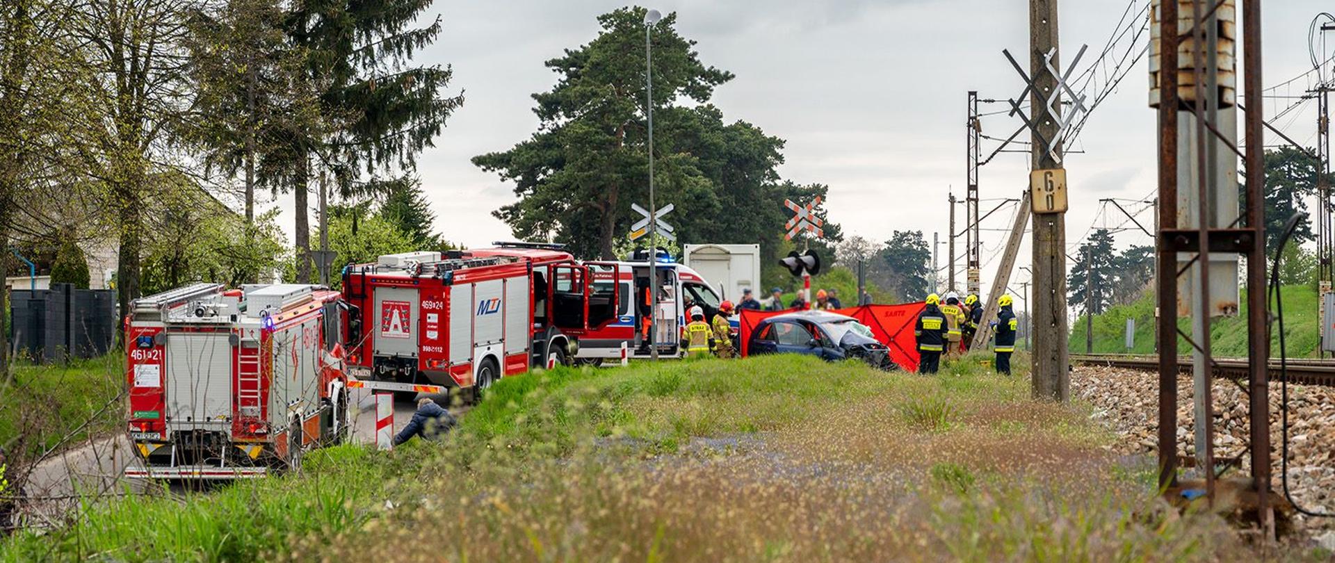 Na zdjęciu widoczny rozbity samochód osobowy przy na przejeździe kolejowym, obok samochody pożarnicze na jezdni