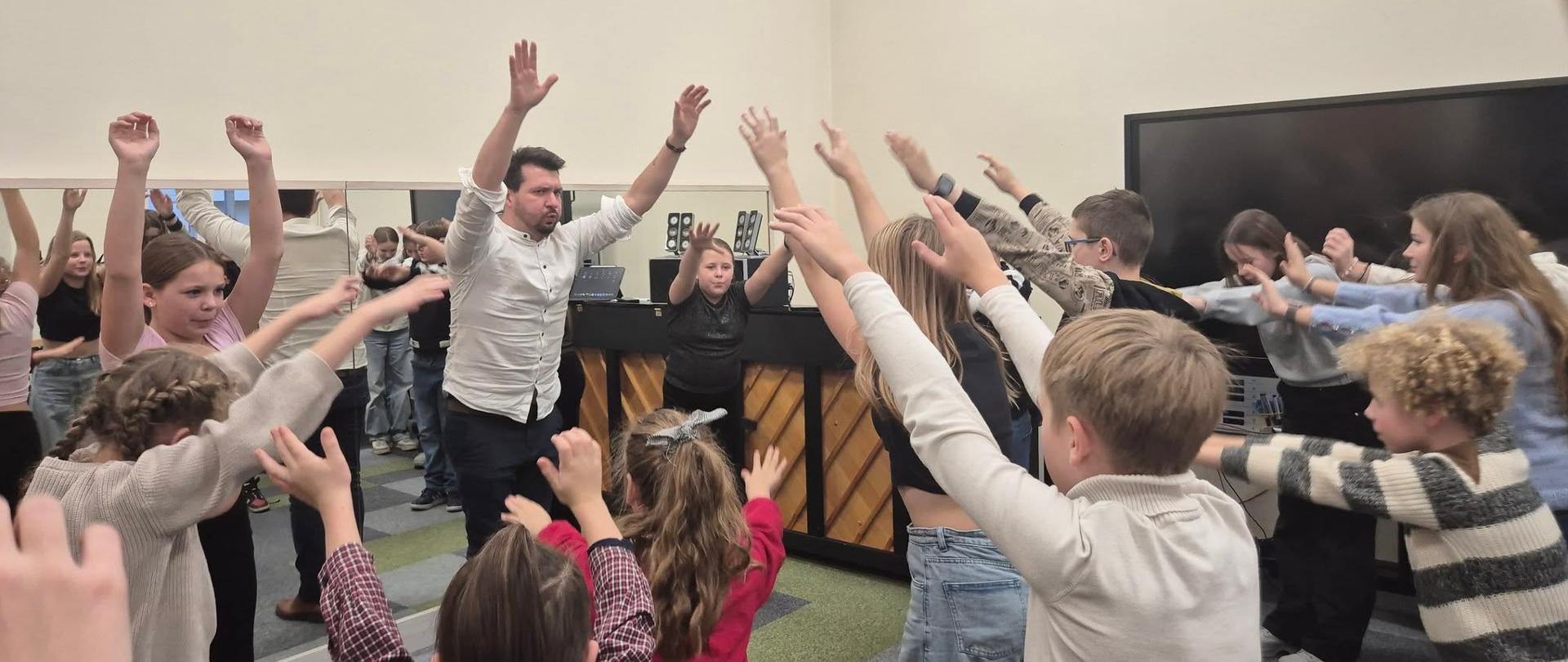 Na zdjęciu uczniowie z podniesionymi rękoma wykonują ćwiczenia oddechowe pod okiem prowadzącego wykładowcy.