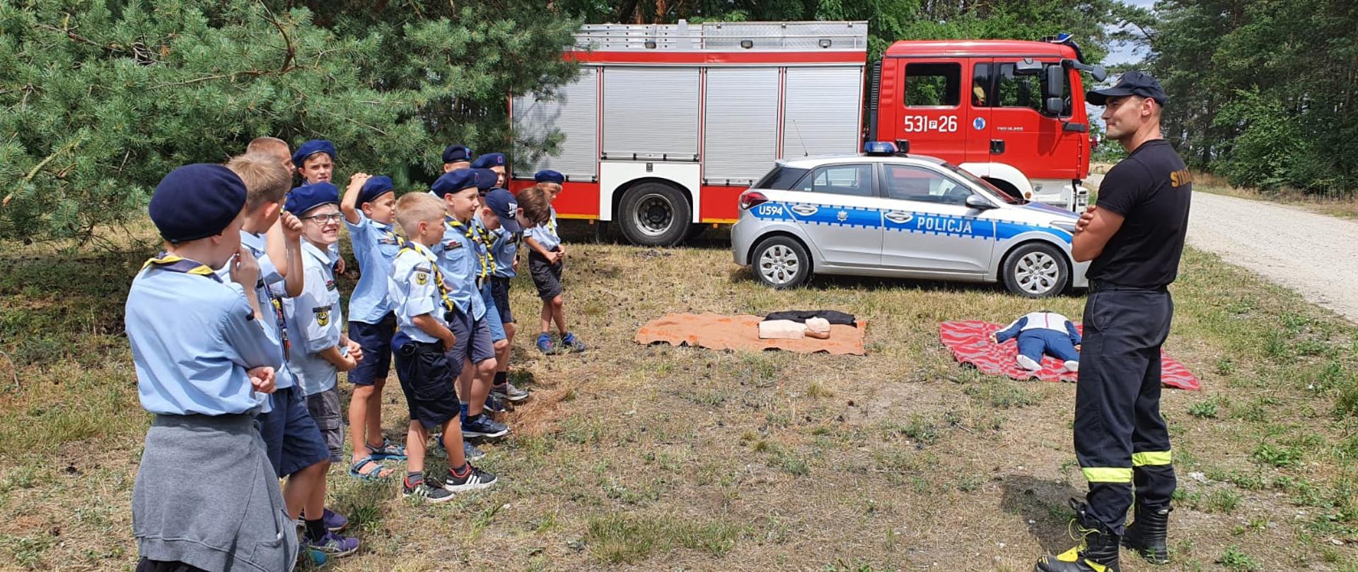Harcerze ustawieni w szeregu, na przeciwko strażak, który udziela pogadanki, W tle las, fantomy, radiowóz i samochód strażacki. Całość zlokalizowana na polanie leśnej.