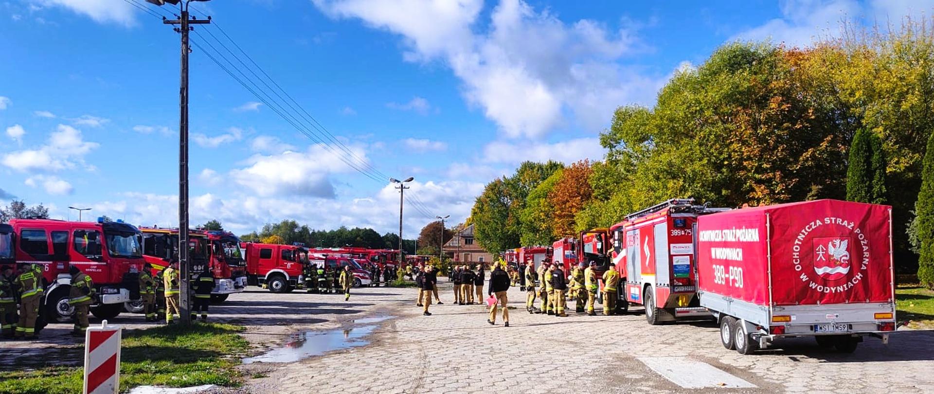 Na zdjęciu widoczny jest plac, na którym przygotowano siły i środki do rozpoczęcia ćwiczeń. Po lewej stronie stoją czerwone samochody pożarnicze i przyczepa, a w środkowej części widać strażaków w ubraniach specjalnych, przemieszczających się pomiędzy pojazdami. Po prawej stronie ustawiono kolejne pojazdy ratownicze. W tle znajdują się drzewa, słupy oświetleniowe oraz niebieskie niebo z chmurami.