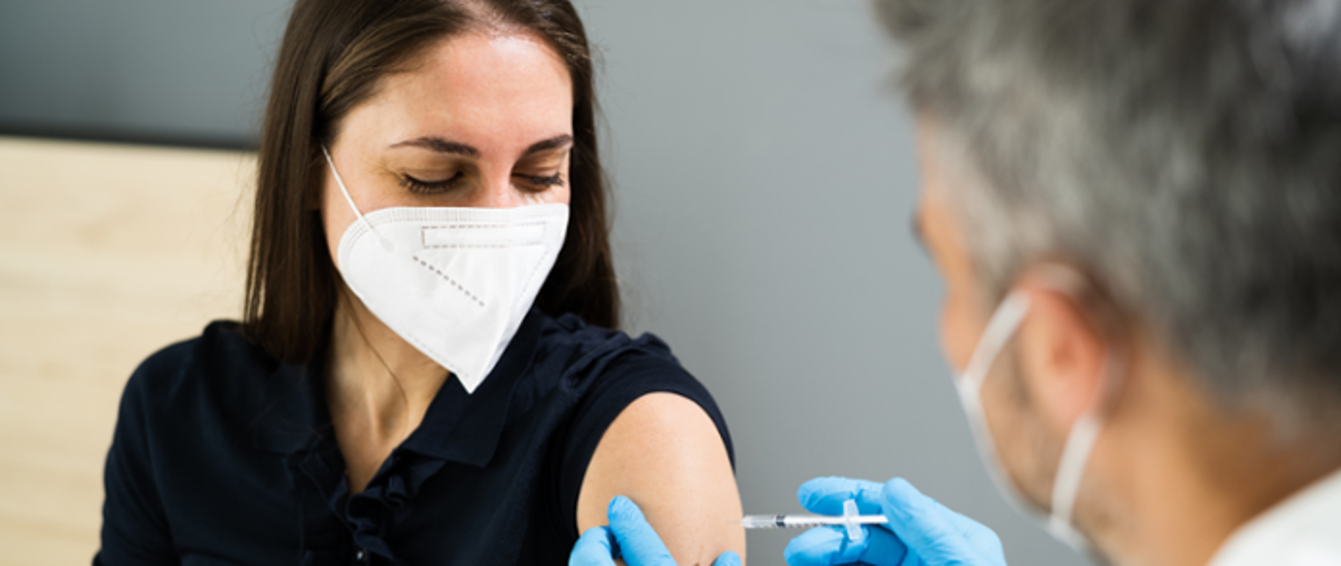 Młoda kobieta ubrana w czarną bluzkę i białą maseczkę przyjmuje szczepionkę w ramię. 
