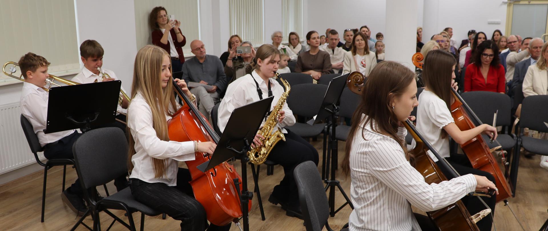 W sali na krzesłach siedzą uczniowie szkoły muzycznej. Trzy uczennice grają na wiolonczelach, trzech uczniów gra na instrumentach dętych: saksofonie, trąbce i puzonie. W tle publiczność słuchająca koncertu