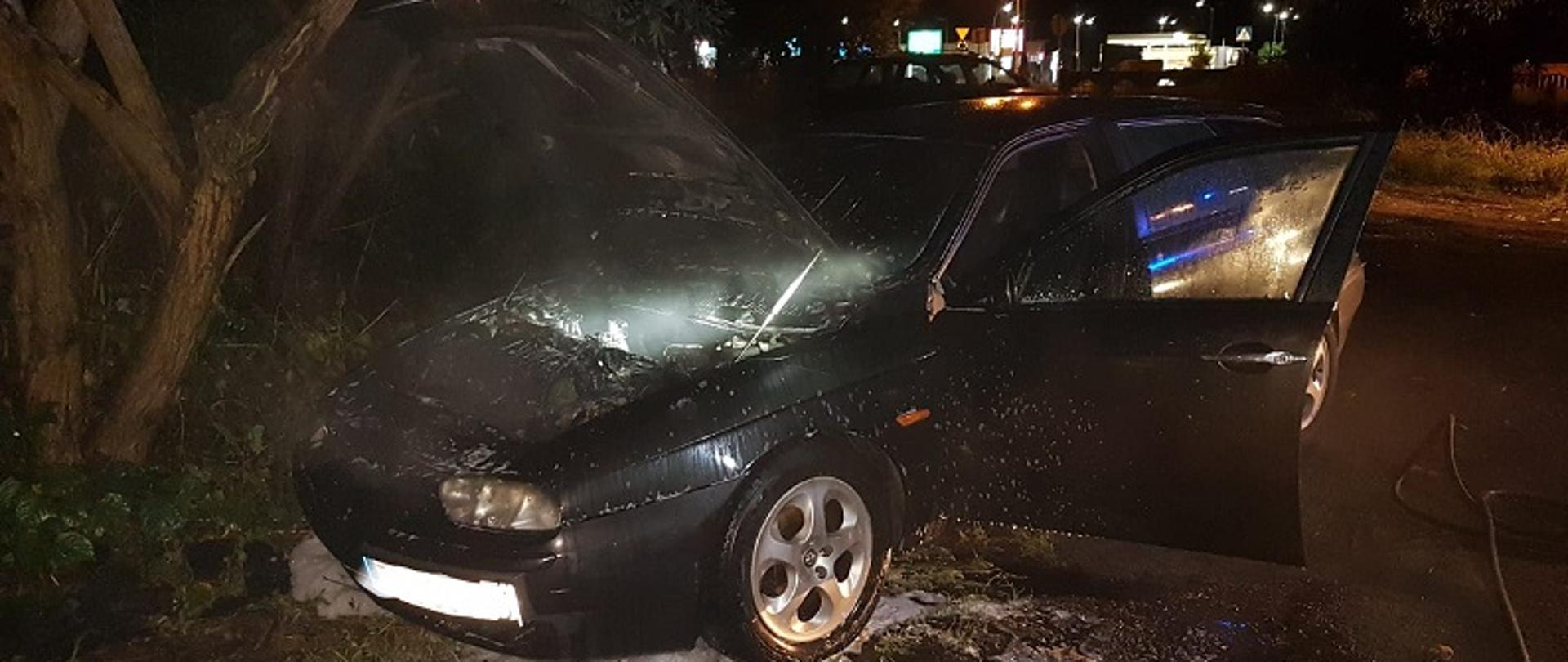 Zdjęcie przedstawia od przodu samochód osobowy Alfa Romeo z otwartą maską komory silnika. Z wnętrza komory silnika wydobywa się szary dym. Drzwi od strony kierowcy są otwarte. Zdjęcie wykonane nocą. Pod pojazdem pozostałości piany gaśniczej