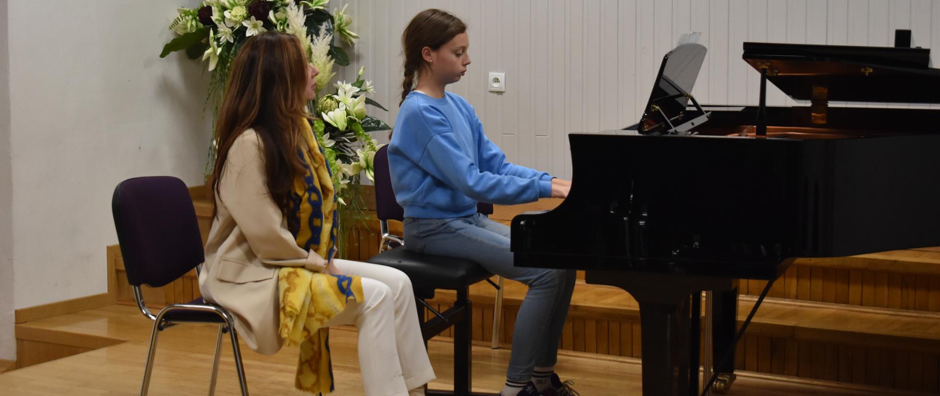 11- Na scenie auli PSM, podczas warsztatów pianistycznych widnieje p. Anna Szajer, która przysłuchuje się grze uczennicy z klasy fortepianu. Kolorystyka zdjęcia jest biało -żółto-zielono-niebiesko-czarna. 