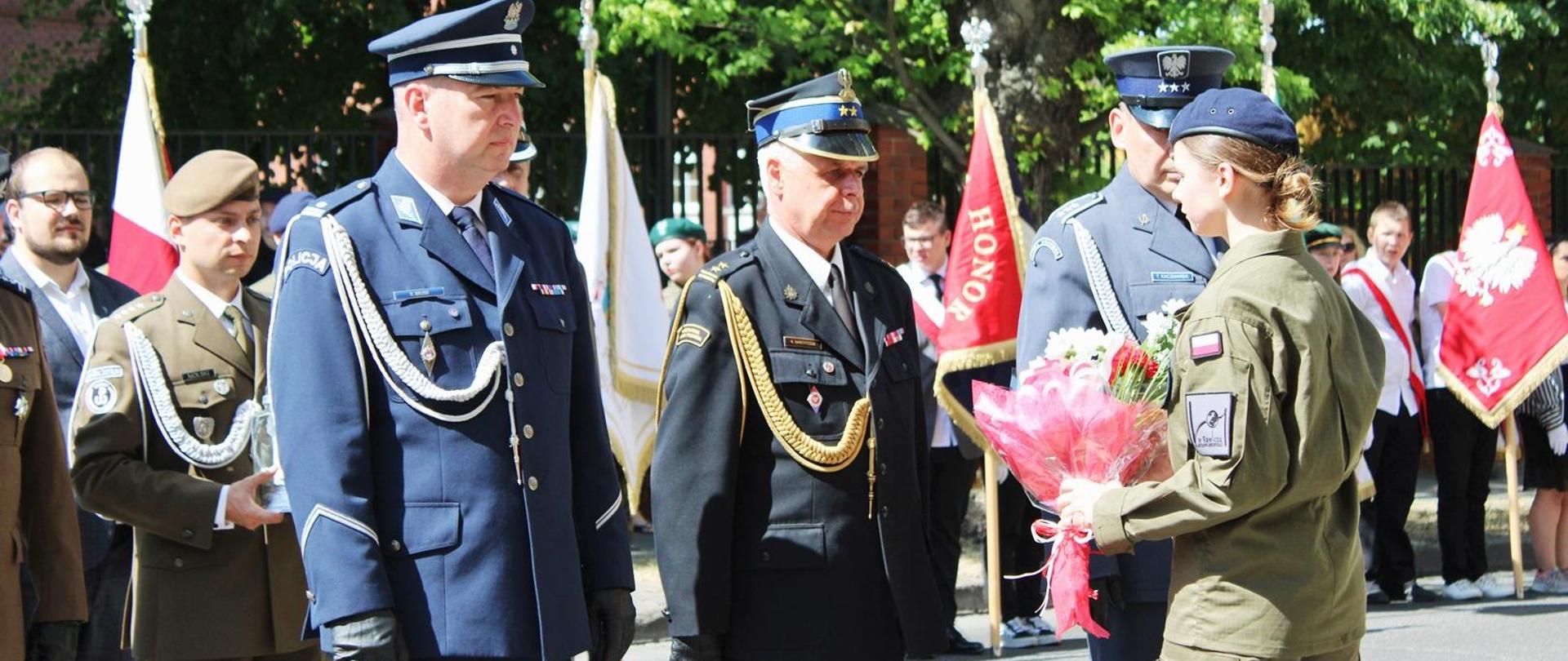 Ulica przed szpitalem w Rawiczu. Delegacja rawickich służb mundurowych (PSP, Policji i SW) w momencie składania wiązanki kwiatów pod tablicą pamiątkową. Strażak przekazał kwiaty uczennicy klasy mundurowej. W tle pozostałe delegacje, poczty sztandarowe oraz drzewa.