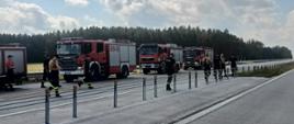 Ustawione na drodze ekspresowej S-61 samochody pożarnicze z JRG PSP Łomża i jednostek OSP z powiatu łomżyńskiego. Strażacy ustawienia są przy barierkach do demontażu. 