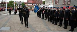 Widok z przodu. Zastępca małopolskiego komendanta wojewódzkiego PSP salutuje, dokonując przeglądu pododdziałów, za nim idzie dowódca uroczystości. Po prawej stronie poczet sztandarowy i pododdział PSP.