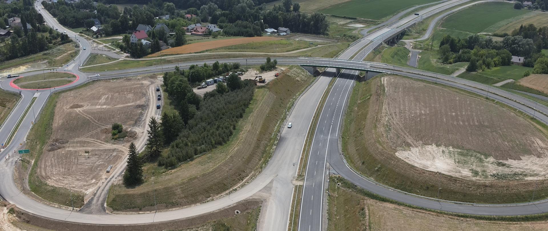 Węzeł DK9 DK74 - zdjęcie wykonane z drona, dwujezdniowa droga, nad nią wiadukt, od wiaduktu odchodzą łącznice węzła, ta z lewej strony dochodzi do ronda 