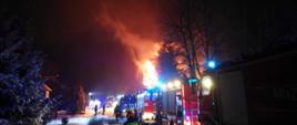 Na zdjęciu w porze nocnej widoczne samochody pożarnicze i strażacy przemieszczający się do pożaru budynku mieszkalnego. Warunki zimowe, śnieg, strażacy w trakcie rozwijania linii gaśniczych.
