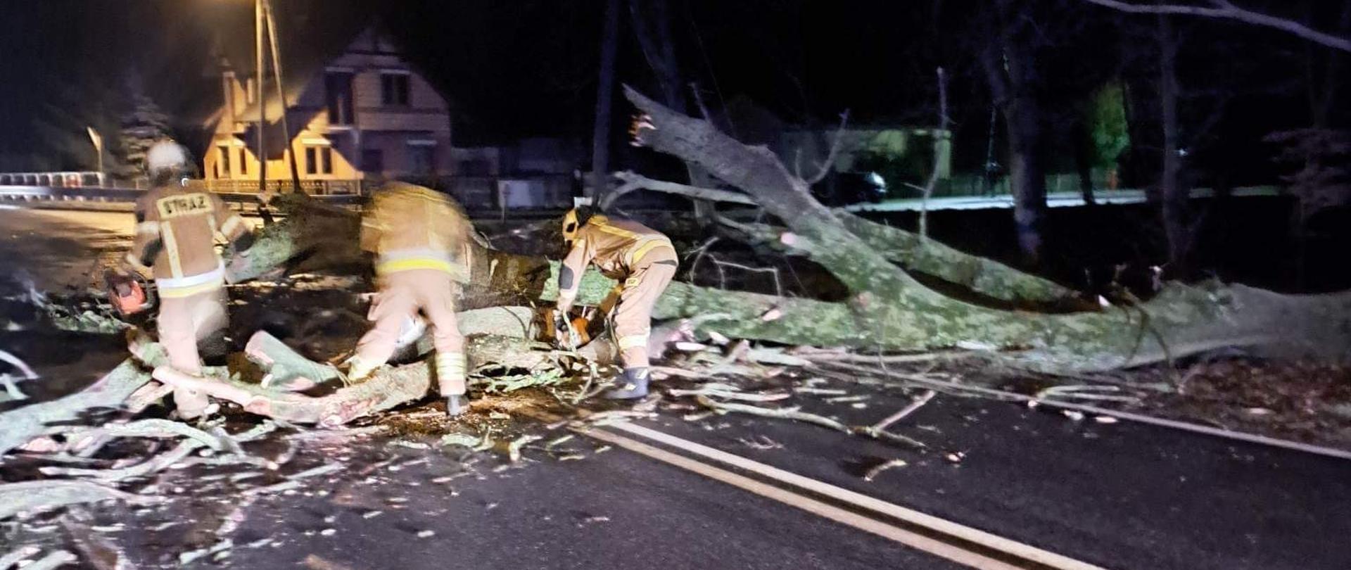 drzewo lezy przewrócone na drodze trzech strażaków z pilarkami uprzątają drzewo w oddali widać słup oświetleniowy oraz dom