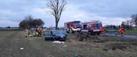 Na zdjęciu rozbity samochód obok strażacy udzielający pomocy osobie poszkodowanej na polu, z boku ratownicy medyczni z noszami przy samochodzie ZRM na jezdni, za nimi samochód pożarniczy