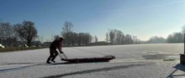 Trwają ćwiczenia z zakresu ratownictwa lodowego. Wyznaczony ratownik za pomocą sani lodowych porusza się po lodzie do miejsca w którym znajduje się osoba poszkodowana. Strażak ubrany w kombinezon do pracy w wodzie, kask , zabezpieczony linią asekuracyjną, 