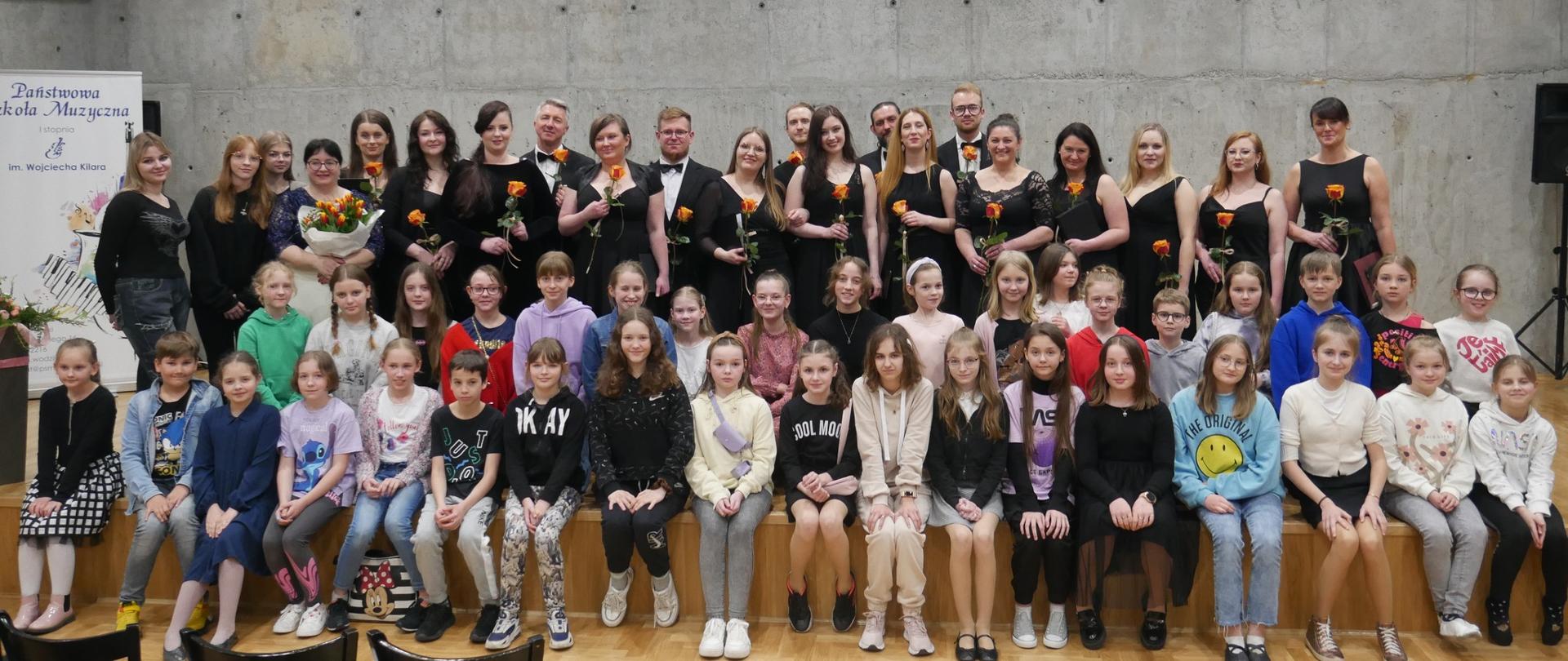 Duża grupa uczniów oraz chórzyści Chóru Kameralnego Ad Libitum na tle sceny sali koncertowej szkoły muzycznej