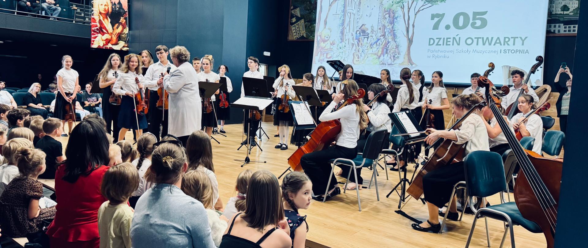 Koncert orkiestry smyczkowej w nowoczesnej sali koncertowej z okazji Dnia Otwartego (7.05). Na scenie młodzi muzycy grają na instrumentach smyczkowych - skrzypcach, altówkach, wiolonczelach i kontrabasach, pod kierunkiem dyrygenta. W tle wyświetlana jest prezentacja multimedialna z napisem "7.05 DZIEŃ OTWARTY" i ilustracją przedstawiającą dzieci grające na instrumentach. Publiczność siedzi na drewnianych ławkach i podłodze, obserwując występ. Sala ma nowoczesną architekturę z ciemnymi ścianami, profesjonalnym oświetleniem scenicznym i dużym ekranem projekcyjnym. 
