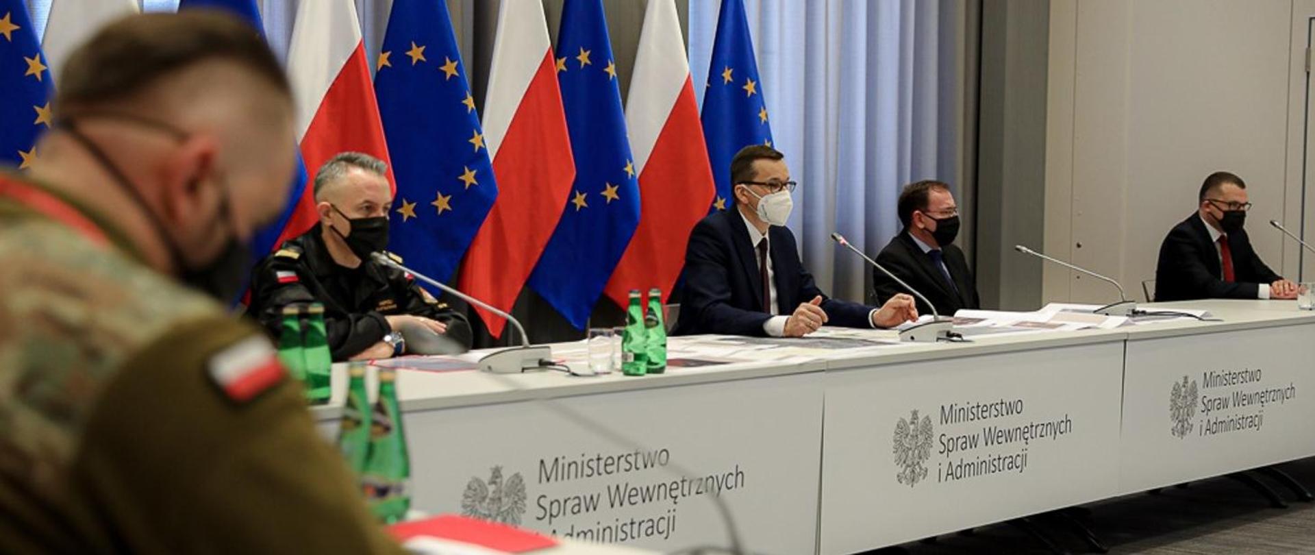 Zdjęcie przedstawia posiedzenie sztabu kryzysowego w sprawie sytuacji powodziowej w Polsce w MSWiA. Na zdjęciu widoczni są siedzący: w środku premier Mateusz Morawiecki, po prawej stronie minister spraw wewnętrznych i administracji Mariusz Kamiński, po lewej stronie komendant główny Państwowej Straży Pożarnej nadbryg. Andrzej Bartkowiak. W tle flagi państwowe i Unii Europejskiej.