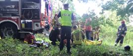 Na zdjęciu strażacy udzielający kwalifikowanej pomocy dla poszkodowanego kierowcy, w tle policjant i osoby postronne.