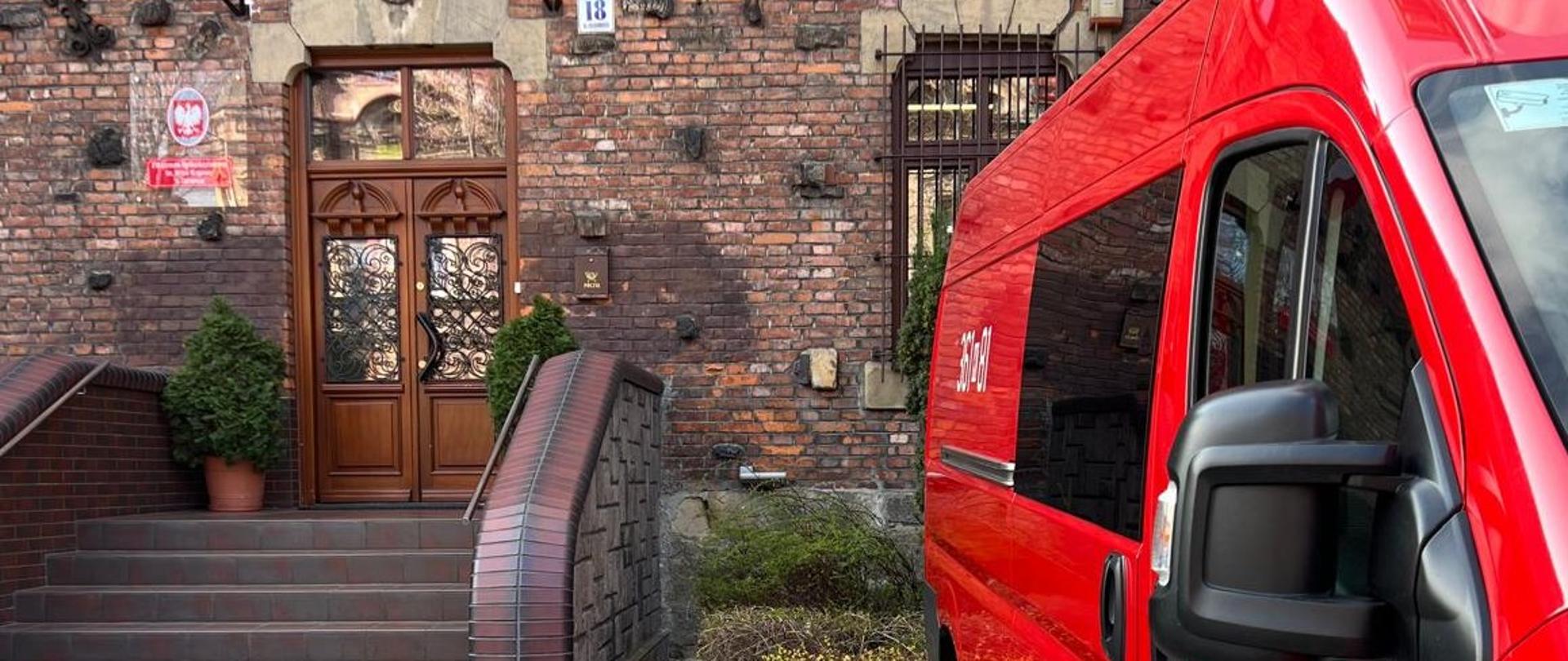 Na kostce przed budynkiem szkoły stoi czerwony samochód strażacki. Do wejścia szkoły prowadzą schody nad wejściem wiszą flagi Polski