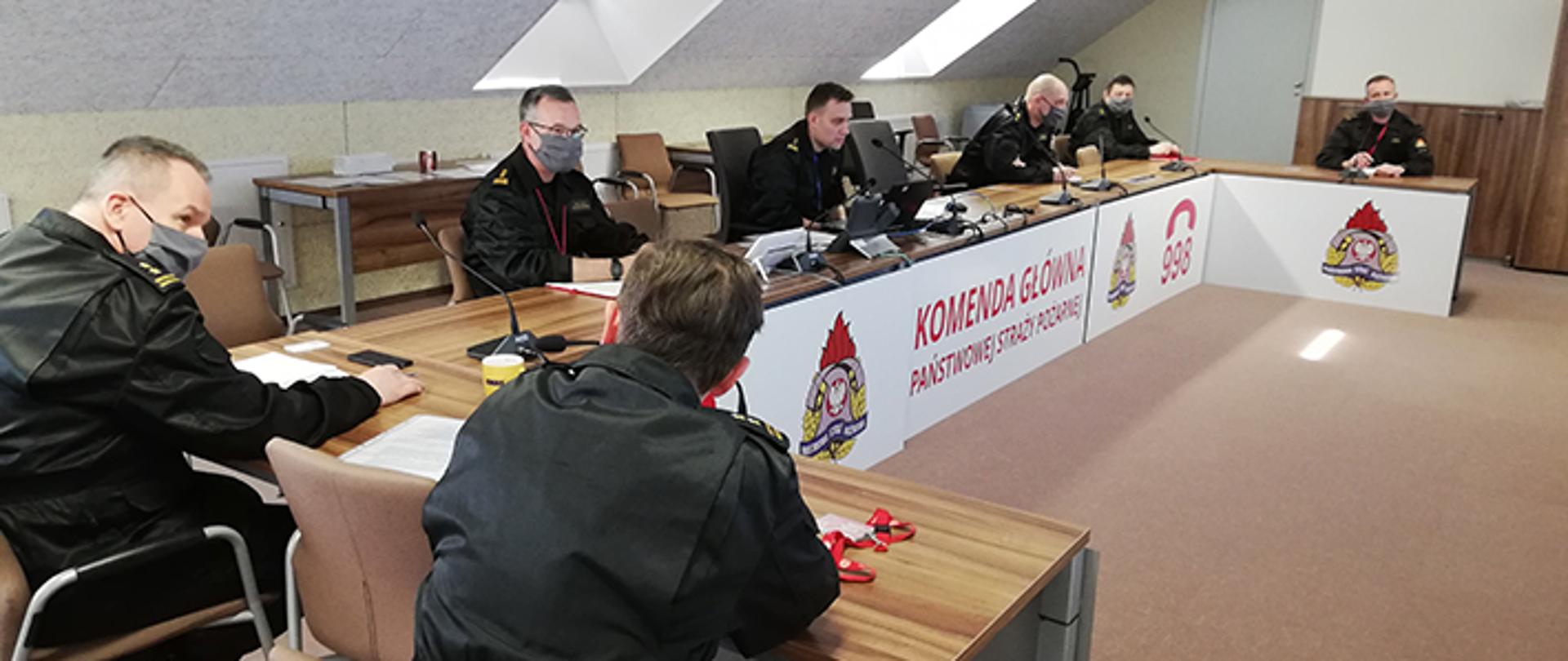 Strażacy w mundurach dowódczo-sztabowych, siedzący przy stole podczas wideokonferencji