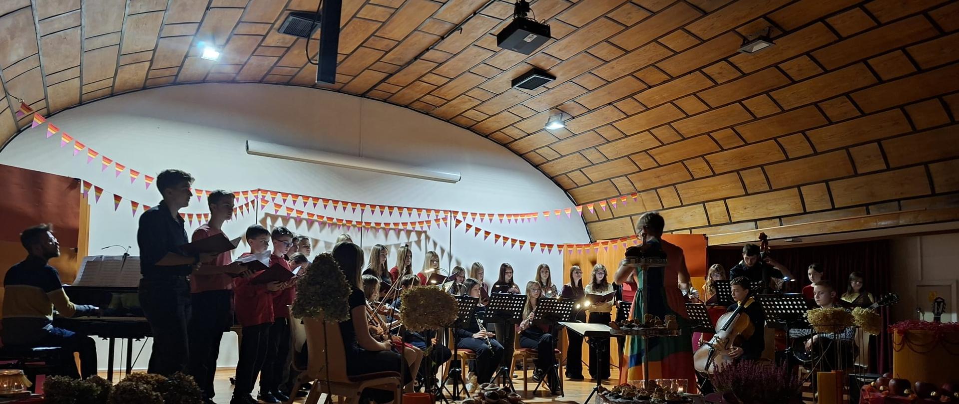 Duże grupa młodzieży na szkolnej scenie gra koncert.